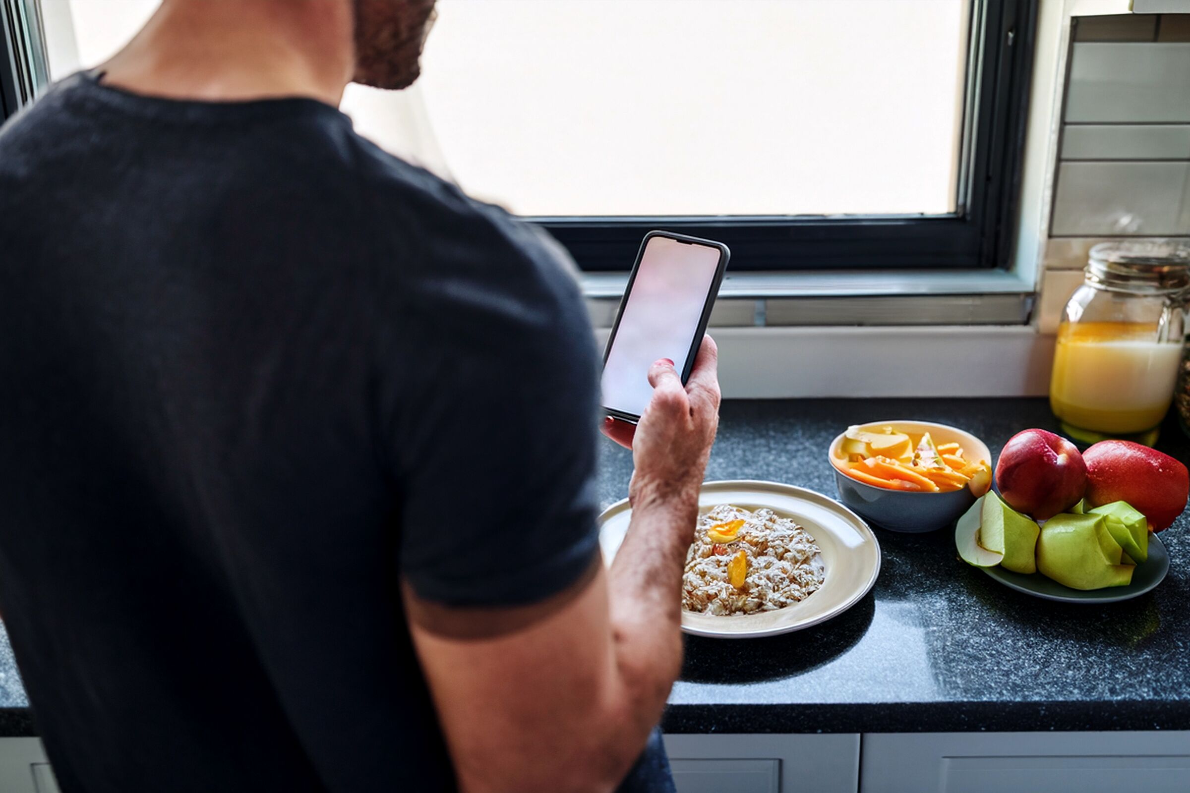 A person at a kitchen counter in morning light, phone in hand, a plate of breakfast in front of them